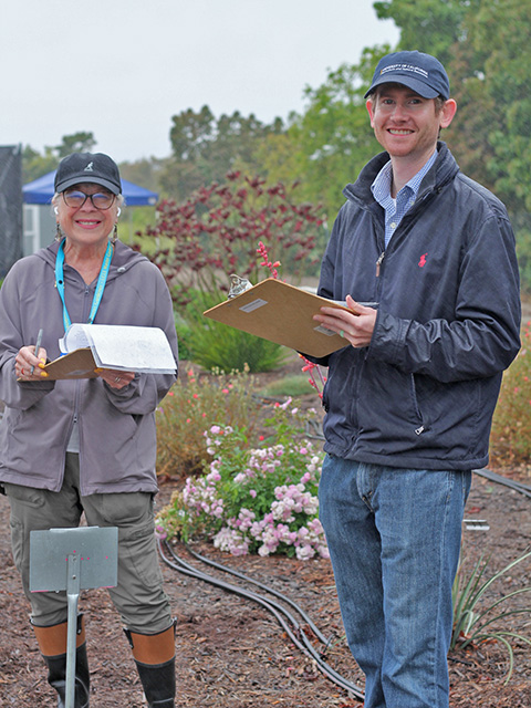 A man and a woman standing outdoors, holding clipboards in their hands, with a garden behind them.
