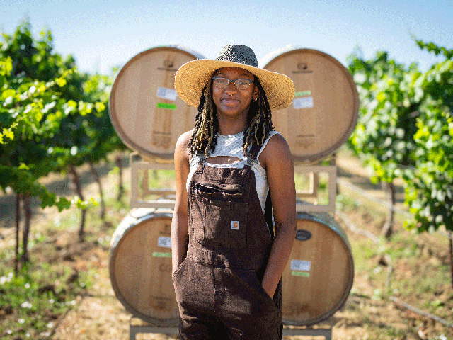 Young African-American woman standing in front of wooden wine barrels set between rows of grapevines. She is wearing a straw hat and brown overalls.