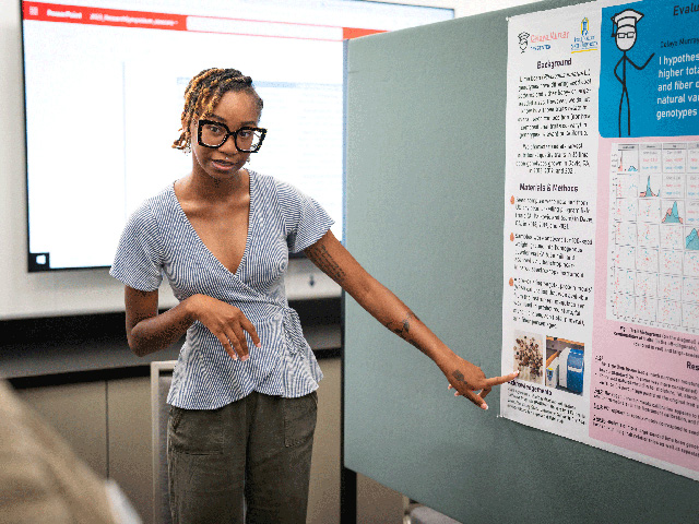A young, African-American woman points to a poster while addressing an onlooker.