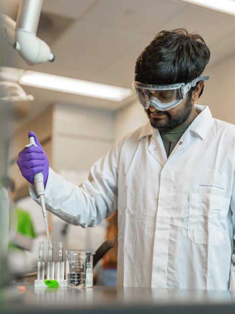 A dark-haired young man wearing a white lab coat and plastic goggles stands in a lab