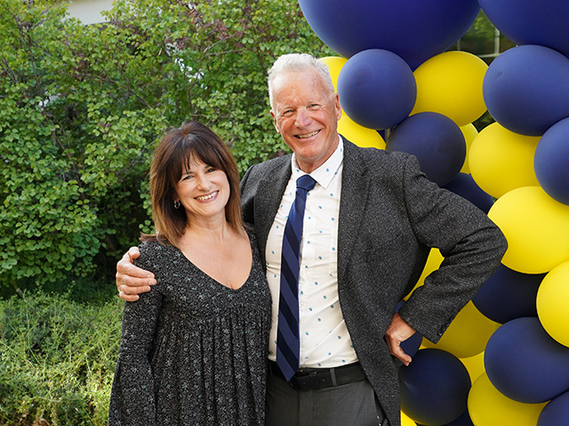A woman and a man, smiling. Yello and blue baloons are next to them.