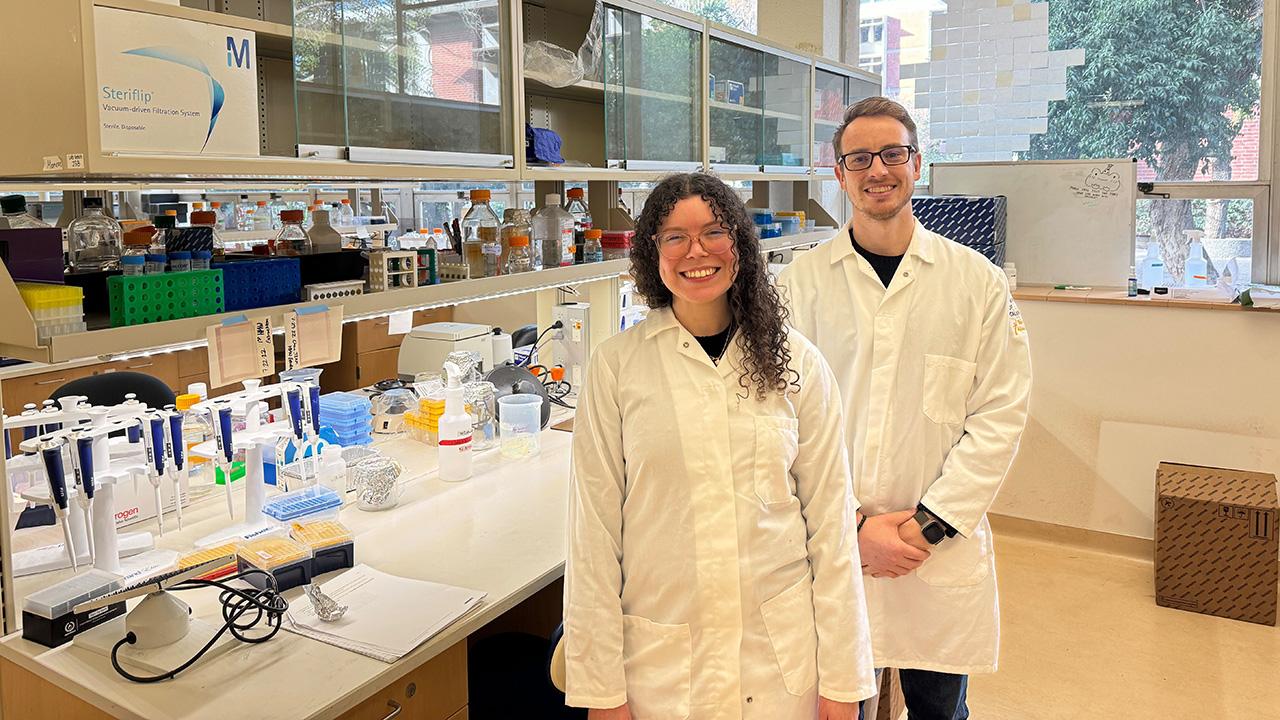 A woman and a man, wearing white lab coats and standing in a lab