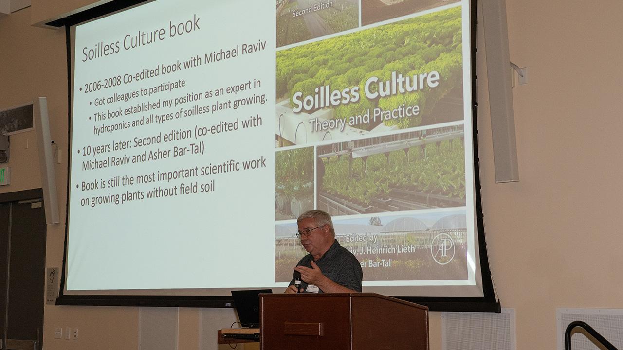 A man speaking, with a projector screen above him showing the words: “Soilless Culture book.”
