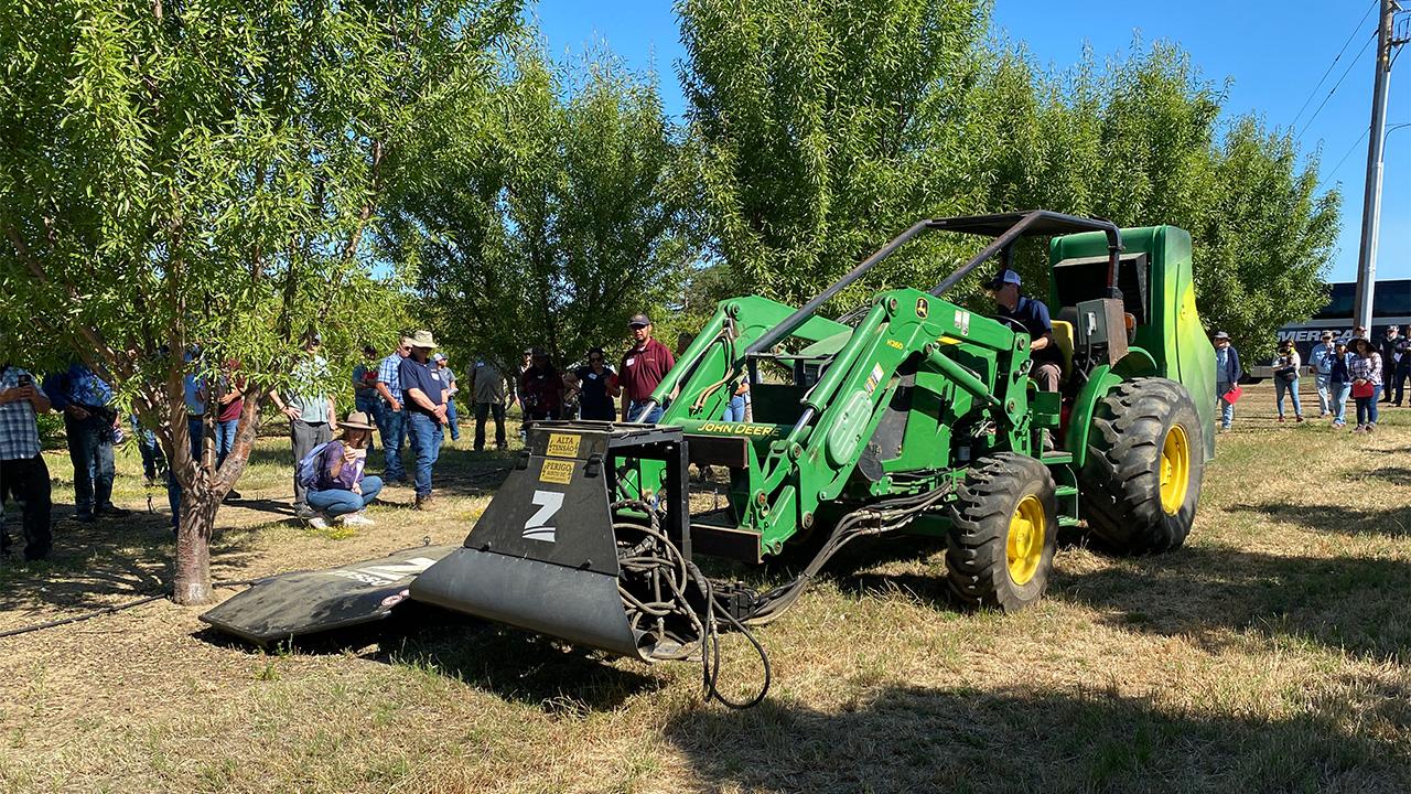 Large green tractor-like machine with an arm extended to the right side, at the edge of a row of green-leafed trees in an orchard. People standing on either side, watching.